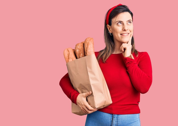 Jeune belle femme tenant un sac en papier avec du pain visage sérieux pensant à la question avec la main sur le menton réfléchie à l'idée confuse