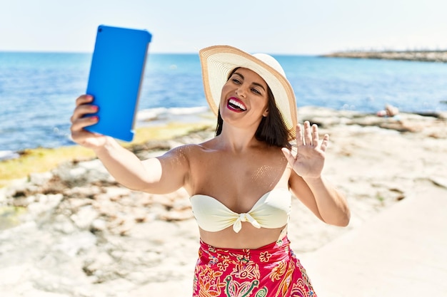 Jeune belle femme touriste hispanique souriant confiant ayant un appel vidéo au bord de la mer