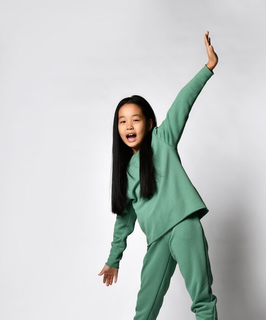 Jeune belle fille asiatique active aux cheveux longs dans un sweat-shirt vert moderne de couleur verte, le sweat-shirt écarte les bras comme des ailes et se réjouit