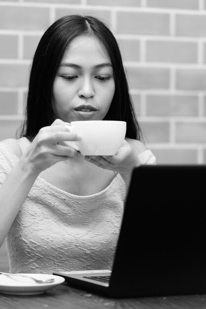 Jeune belle fille asiatique buvant du café avec les deux mains avec un ordinateur portable sur une table en bois sur un mur de briques