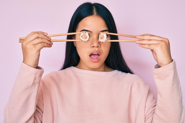 Jeune belle fille asiatique mettant des sushis sur les yeux à l'aide de baguettes en face de choc sceptique et sarcastique surpris avec la bouche ouverte