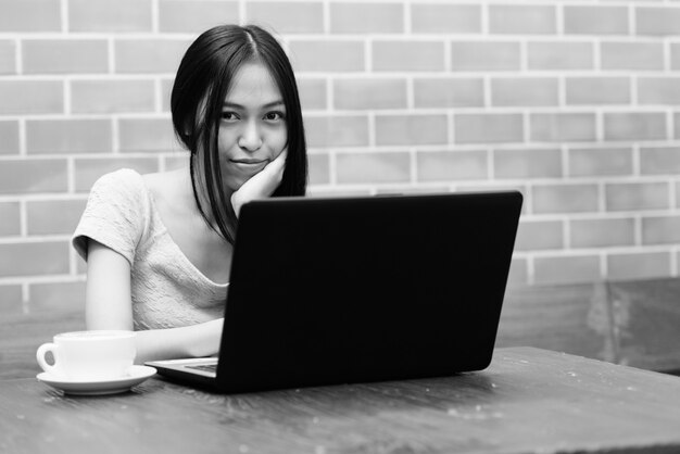 Jeune belle fille asiatique reposant le menton à portée de main avec un ordinateur portable et un cappuccino sur une table en bois sur un mur de briques