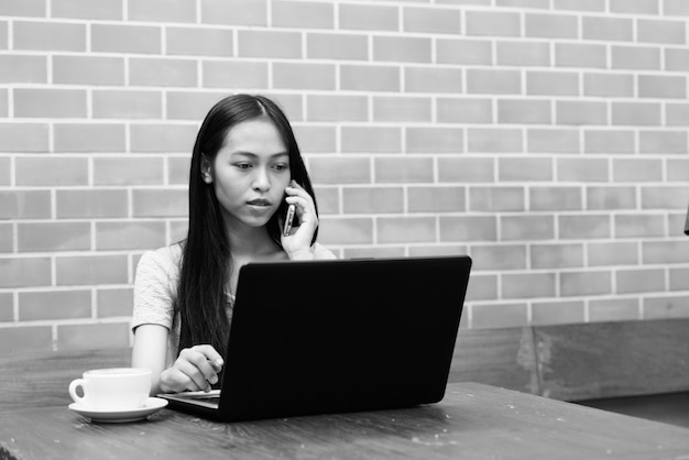 Jeune Belle Fille Asiatique Utilisant Un Ordinateur Portable Sur Une Table En Bois Tout En Parlant Au Téléphone Portable Sur Un Mur De Briques