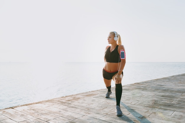 La jeune belle fille athlétique aux longs cheveux blonds dans les écouteurs et le téléphone portable sur sa main écoutant de la musique et faisant des étirements au lever du soleil sur la mer