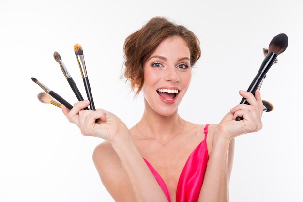 Jeune belle fille aux cheveux bruns ondulés, peau propre, dents plates, beau sourire, en jersey rose, détient une variété de pinceaux de maquillage