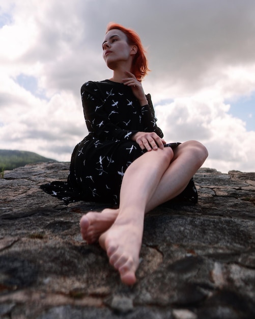 Jeune belle fille aux cheveux rouges est assise sur le rocher dans la forêt et se détend sous un ciel nuageux.