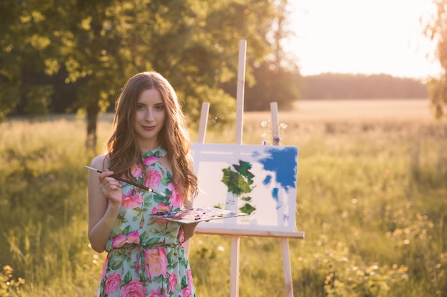 Jeune belle fille aux longs cheveux naturels dessine une image avec un chevalet dans la nature.