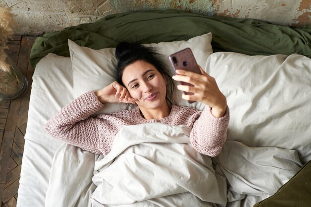 Jeune belle fille bavardant et posant sur le smartphone et riant en position couchée dans son lit à la maison