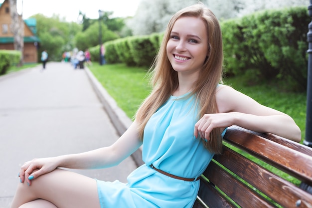 Jeune belle fille blonde assise sur un banc dans le parc et souriant