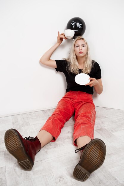 Jeune belle fille blonde avec un ballon noir au chapeau assis sur le sol en buvant du café sur un mur blanc