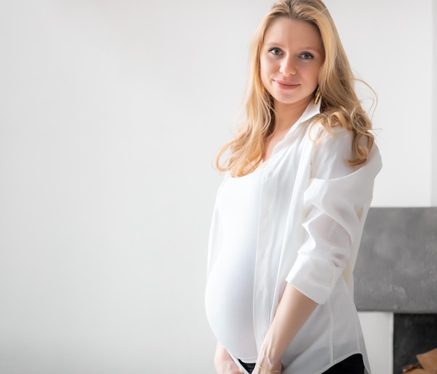 Jeune belle fille blonde caucasienne enceinte souriante heureuse debout dans le salon de la lumière blanche