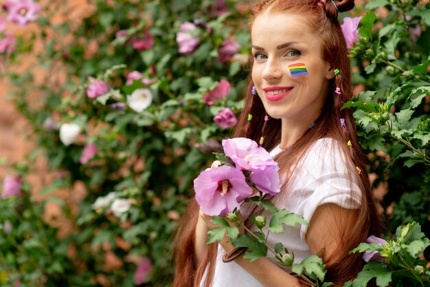 Jeune belle fille brune aux cheveux longs avec arc-en-ciel lgbt sur son visage posant en fleurs épanouies