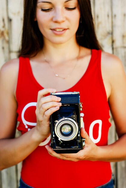 Jeune belle fille brune en short chemise et jeans rouge, posant avec un appareil photo sur le fond en bois rustique.