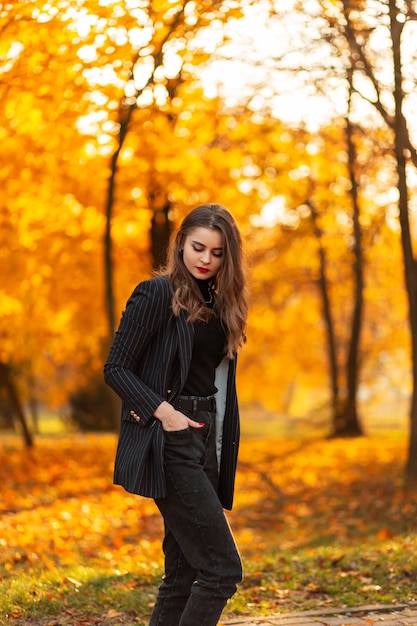 Jeune belle fille dans un costume élégant noir avec un blazer à la mode sur la nature avec des feuilles d'automne lumineuses au coucher du soleil