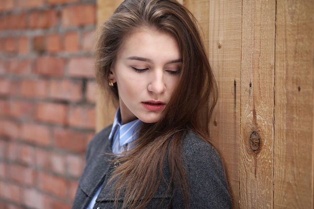 Jeune belle fille dans un manteau près du mur de briques rouges posant