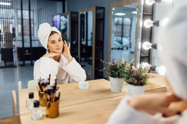 Jeune belle fille dans un peignoir blanc et avec une serviette sur la tête après une douche devant un miroir.