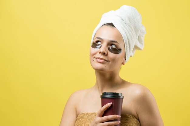 Jeune belle fille dans une serviette blanche sur la tête porte des patchs de gel de collagène sous les yeux Masque sous le visage de traitement des yeux La fille tient un verre de café dans ses mains