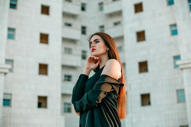 Jeune belle fille dans une veste noire pose près d&#39;une haute maison blanche.