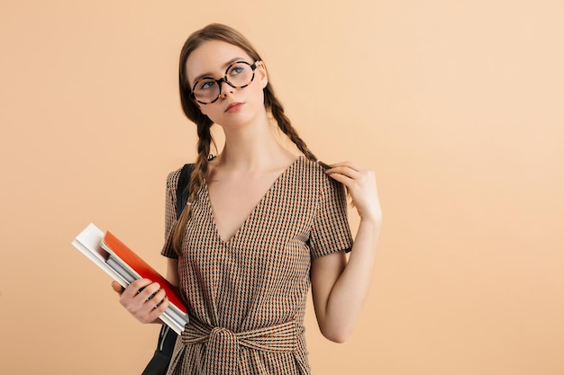 Jeune belle fille avec deux tresses en combinaison de tweed et lunettes avec sac à dos noir sur l'épaule tenant un livre et des blocs-notes à la main tout en regardant rêveusement sur fond beige