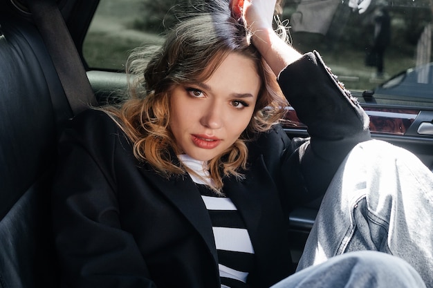 Une jeune et belle fille est assise dans une voiture. Fille élégante dans un grand portrait de costume, dans l'intérieur noir de la voiture.