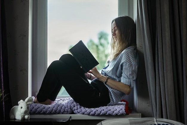 jeune belle fille est assise à la maison sur le rebord de la fenêtre et lit un livre