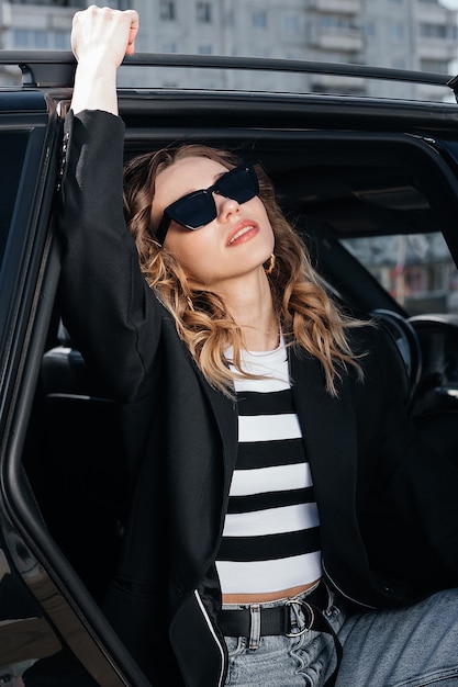 Une jeune et belle fille est assise sur le siège arrière d'une voiture. Une fille élégante en costume et lunettes sort de la voiture.