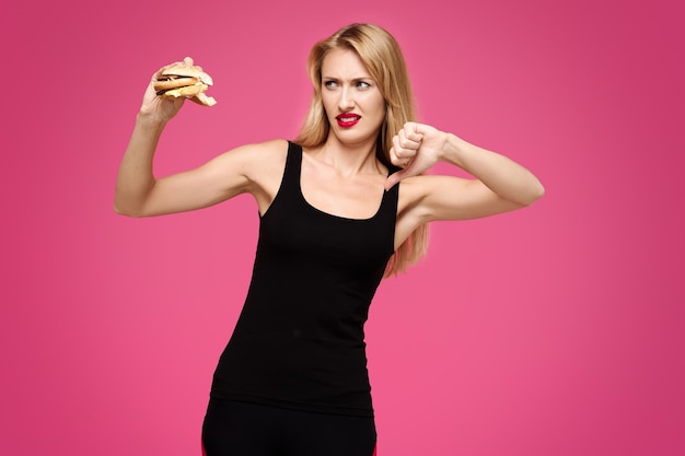 Photo jeune belle fille sur fond rose tient un hamburger dans ses mains et montre son pouce vers le bas