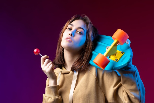 Jeune belle fille hipster à la mode vêtue d'un sweat à capuche tient une planche à roulettes dans ses mains sur un fond de studio violet Mange une sucette