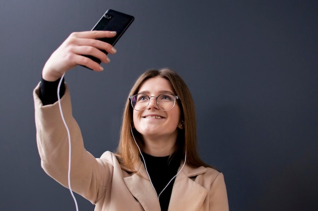 Jeune belle fille à lunettes sur fond sombre écoute de la musique prend un selfie