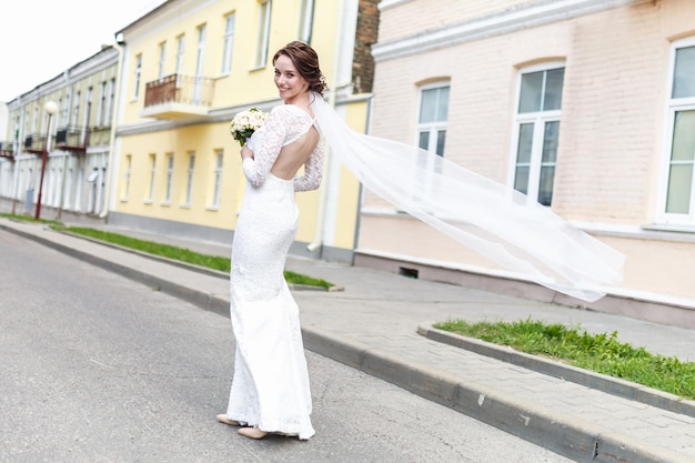 Jeune belle fille mariée avec bouquet de mariée de roses se dresse dans la rue le vent souffle le voile