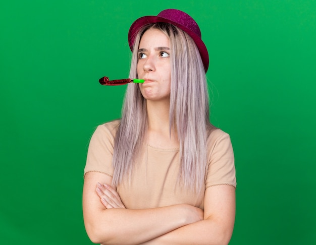 Jeune belle fille mécontente portant un chapeau de fête soufflant un sifflet de fête traversant les mains