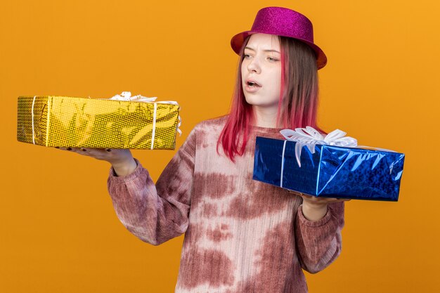 Jeune belle fille mécontente portant un chapeau de fête tenant et regardant des coffrets cadeaux isolés sur un mur orange