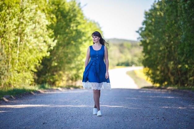 Jeune belle fille modèle grande taille heureuse en robe bleue sur la route goudronnée rurale regardant au loin en été