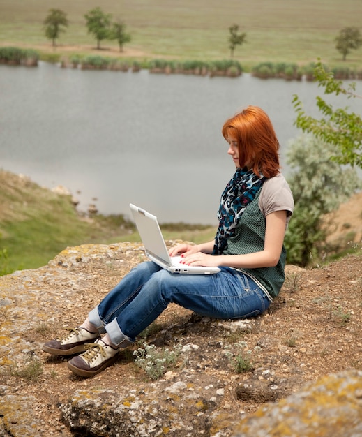 Jeune belle fille avec ordinateur portable au rocher près du lac et de l'arbre.