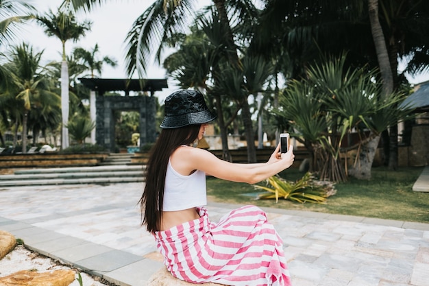 Jeune belle fille en panama noir fait selfie sur un smartphone au bord de la mer dans un parc de loisirs. Vacances sous les tropiques.