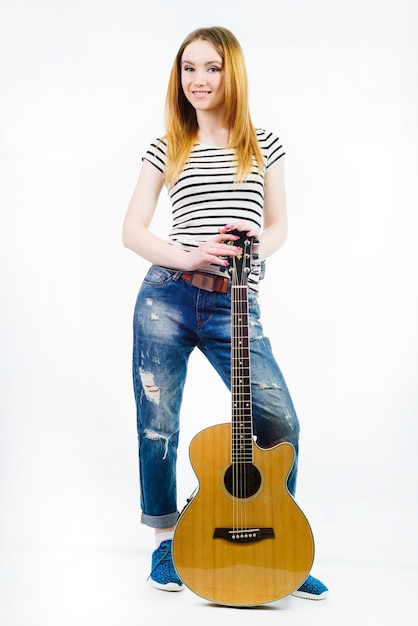 Jeune belle fille portant des jeans et un t-shirt rayé avec une guitare acoustique sur fond blanc