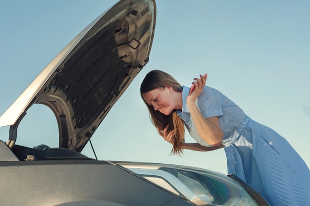 Jeune Et Belle Fille Près D'une Voiture Cassée Avec Un Capot Ouvert. Les Problèmes Avec La Voiture, Ne Démarre Pas, Ne Fonctionne Pas.