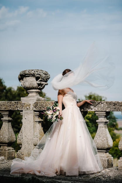 Jeune belle fille en robe élégante est debout et tenant la main bouquet de fleurs roses pastel et de verts près du vieux mur à la nature Le vent jette un voile de mariée à l'extérieur