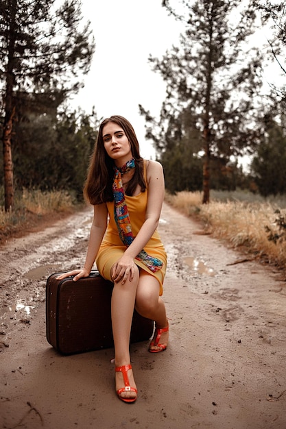 Jeune belle fille en robe jaune avec écharpe colorée assise sur une valise vintage sur une route boueuse trempée après la pluie