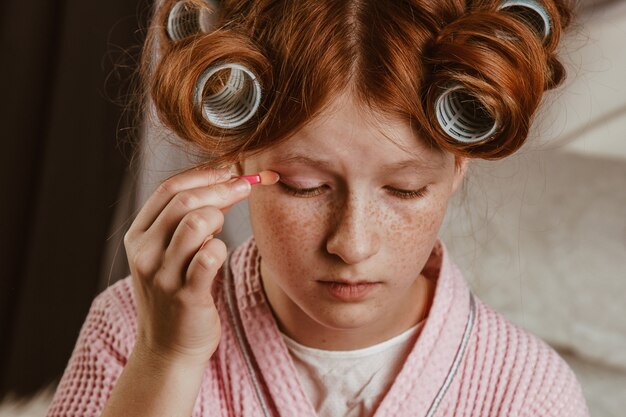 Jeune belle fille se maquille sur le lit dans la chambre. Rousse et calfeutrant avec des bigoudis sur la tête dans un peignoir.