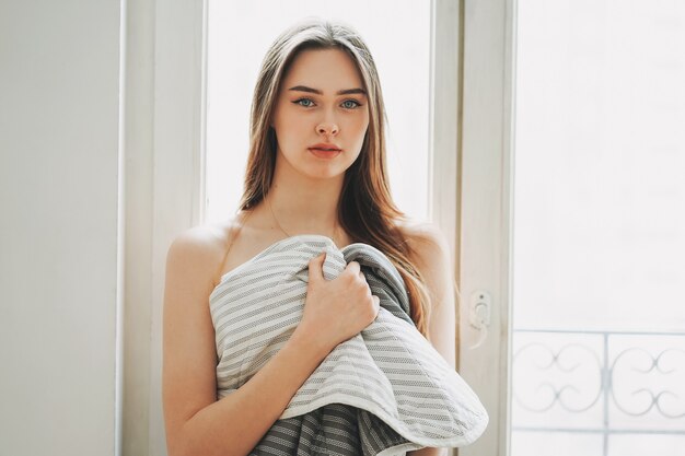 Jeune belle fille sensible aux cheveux longs en couverture en regardant la caméra près de la fenêtre