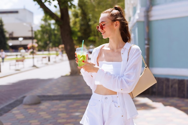 Jeune belle fille en vêtements blancs et verres rouges boit un cocktail rafraîchissant d'été