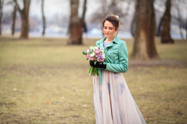 Jeune belle fille vêtue d'une robe longue et d'une veste verte et avec un bouquet de fleurs dans ses mains se promène parmi les arbres du parc. Elle est heureuse et souriante.