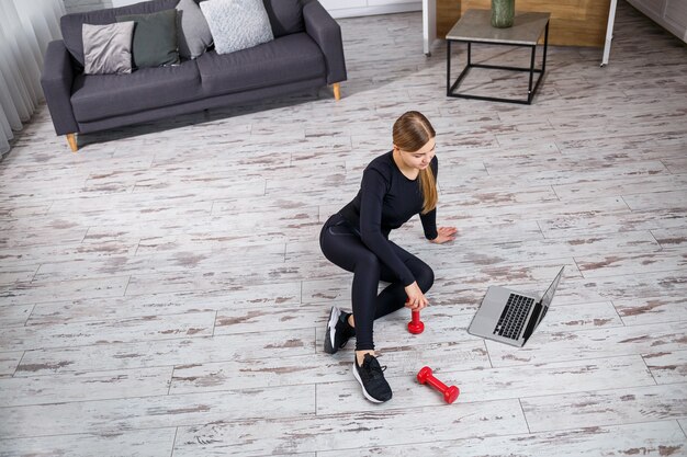 Jeune belle formatrice en haut noir et leggings est assise avec un ordinateur portable et regarde l'entraînement en ligne, le fitness à la maison pendant la quarantaine. Mode de vie sain. Les haltères sont sur le sol