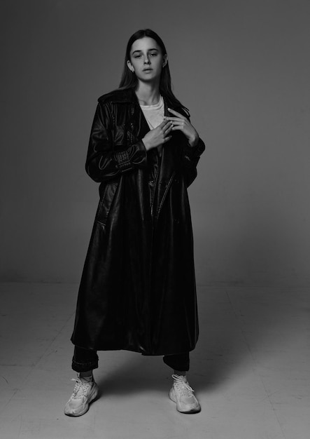 Photo jeune belle mannequin en long manteau en cuir et jeans sombres sur un fond gris foncé