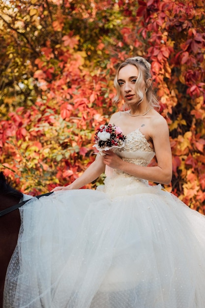 Jeune belle mariée blonde dans une robe blanche duveteuse couvrant une équitation dans la forêt d'automne