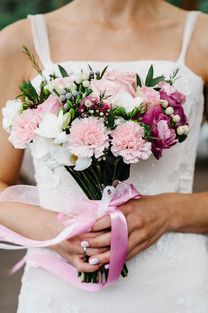 Jeune belle mariée vêtue d'une robe élégante est debout et tient un bouquet de fleurs roses et violettes et de verts avec ruban lors de la cérémonie de mariage