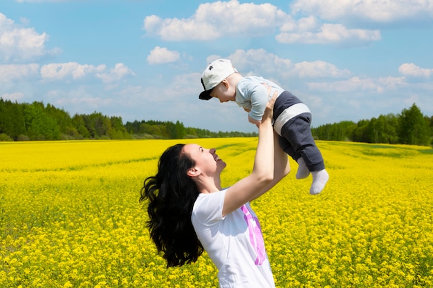 Une jeune et belle mère brune tenant son petit fils dans ses bras dans un champ de fleurs de colza jaune en fleurs.
