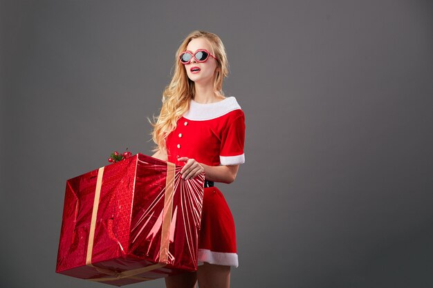 Jeune et belle mrs.Santa Claus en lunettes de soleil vêtue de la robe rouge, des gants blancs et des talons hauts détient l'énorme cadeau de Noël sur le fond gris. .