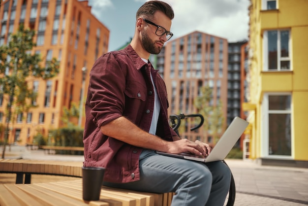 Jeune blogueur bel homme avec du chaume dans des vêtements décontractés et des lunettes travaillant sur un ordinateur portable tout en